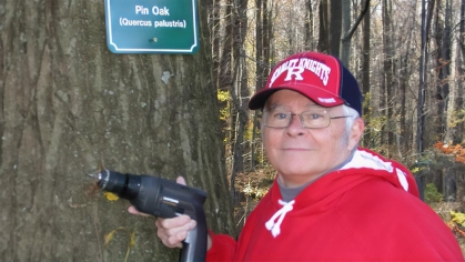 A Master Tree Steward holding an electric drill next to a tree that has a new sign on it that says 'Pin Oak (Quercus palustris)'.